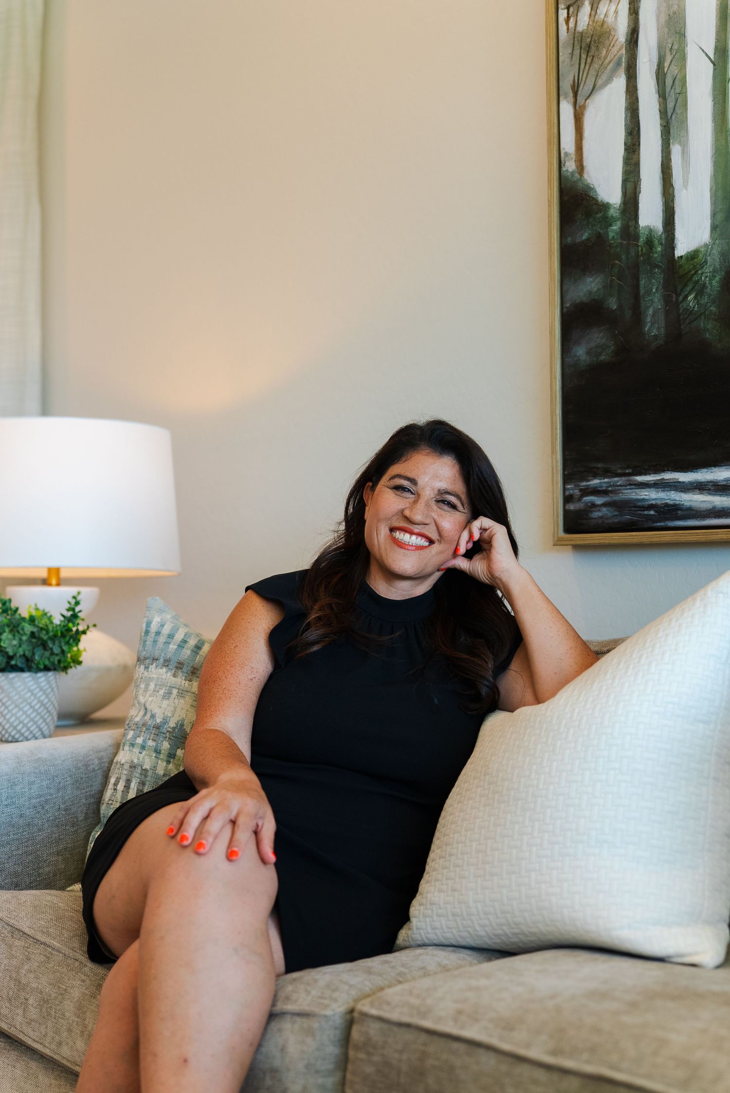 Smiling woman in black dress sits relaxed on gray sofa beside lamp and abstract wall art in elegant room.