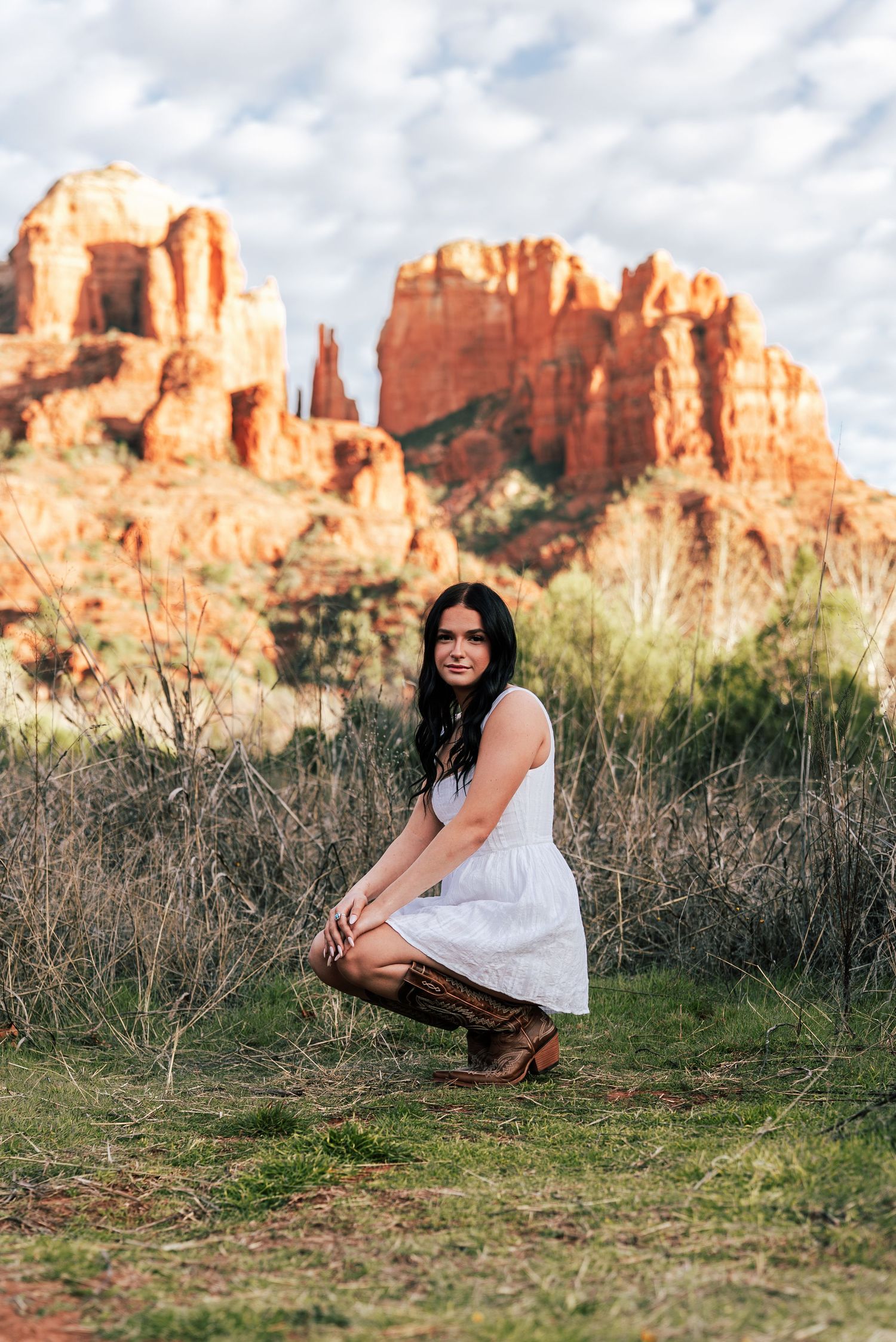 Young woman in white dress crouching on grass with Sedona red rock formations in background.