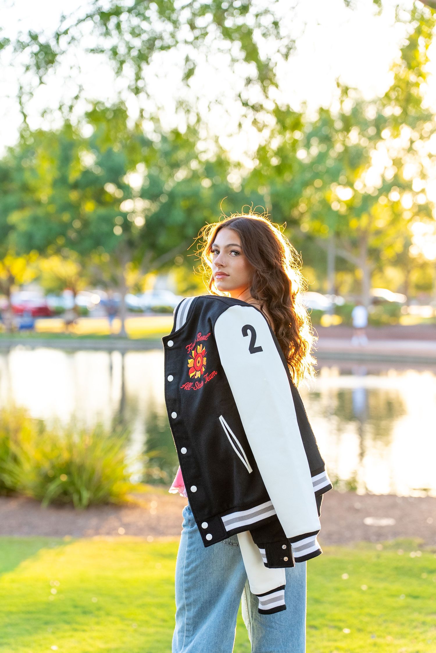 Young woman in black and white varsity jacket with number 2, posing outdoors near a pond in a sunny park.