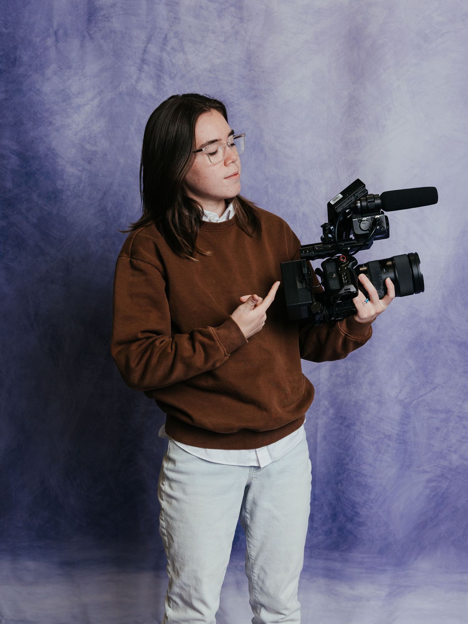 Young person in brown sweater holding a professional video camera against a purple backdrop.