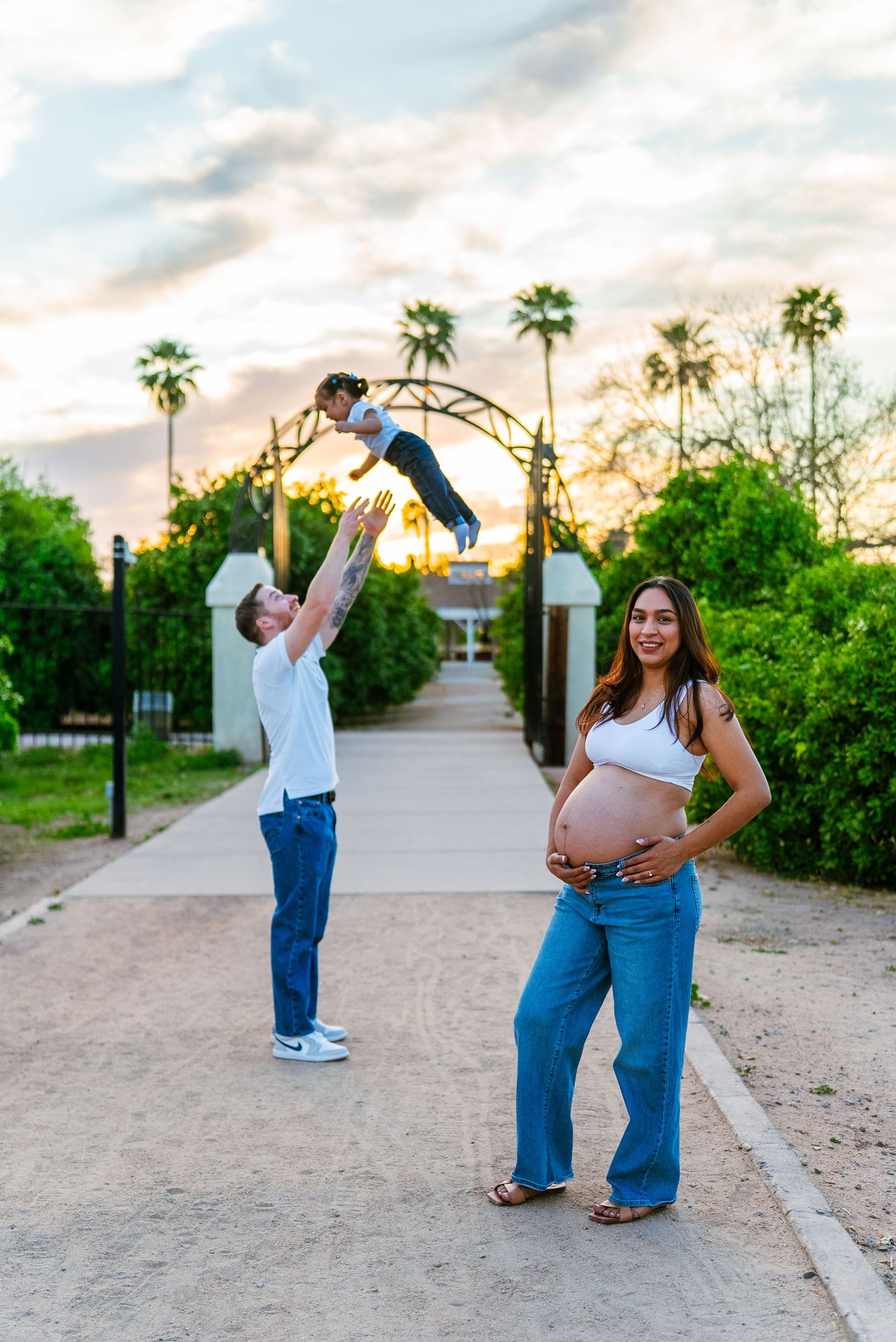 Pregnant woman holds belly while man tosses child in air at sunset park with palm trees.