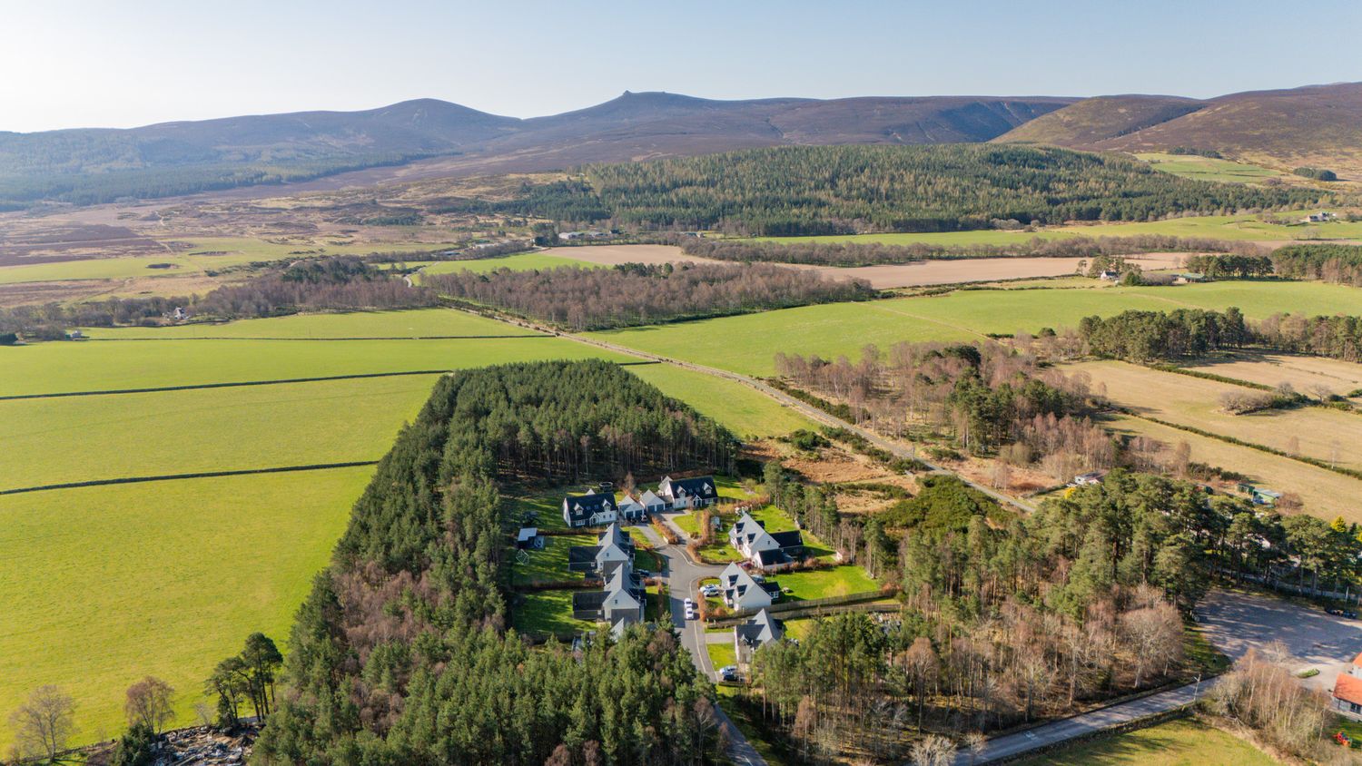 Panoramic drone shot of a secluded estate surrounded by green fields and forested hills.