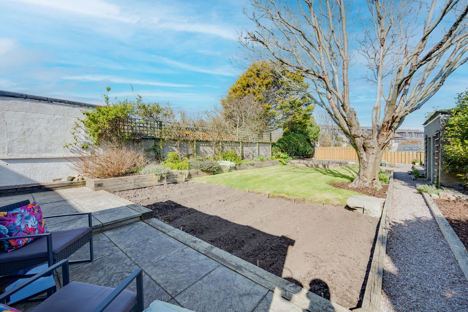 Backyard garden with tilled soil beds, concrete patio, and bare trees against blue sky and wooden fence.