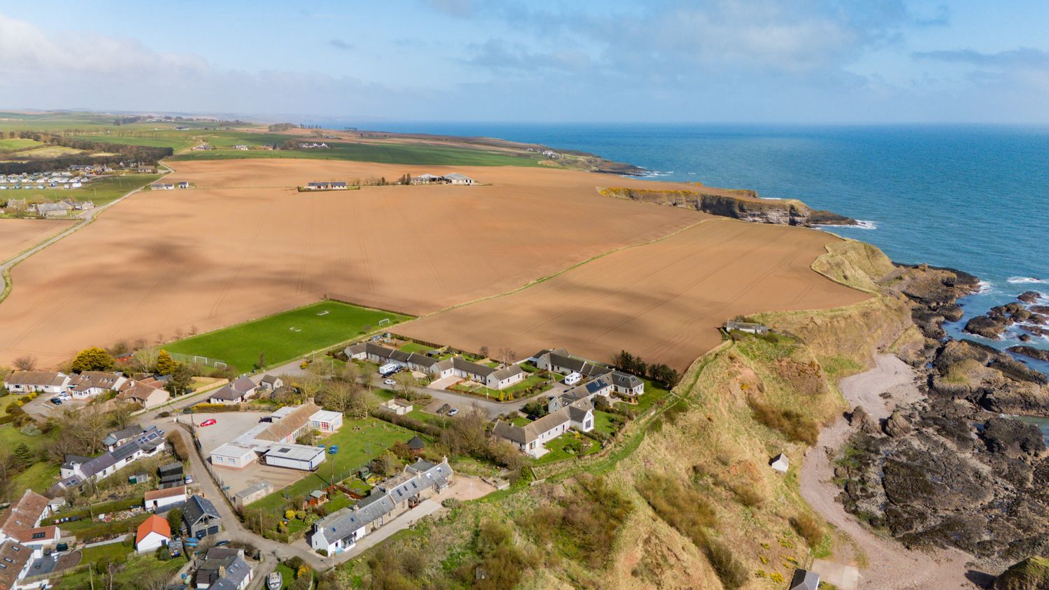 Aerial view of coastal farmland and buildings along rocky cliffs overlooking the blue ocean on a clear day.
