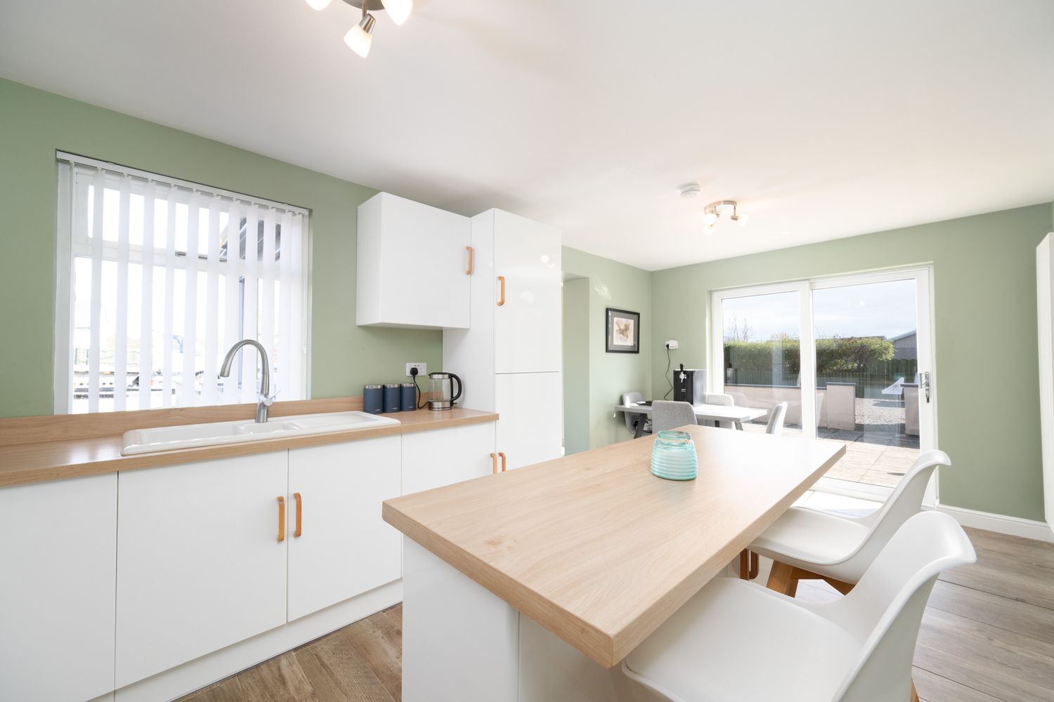 Modern kitchen interior with white cabinets, wood countertops, sage green walls and natural light from large windows.