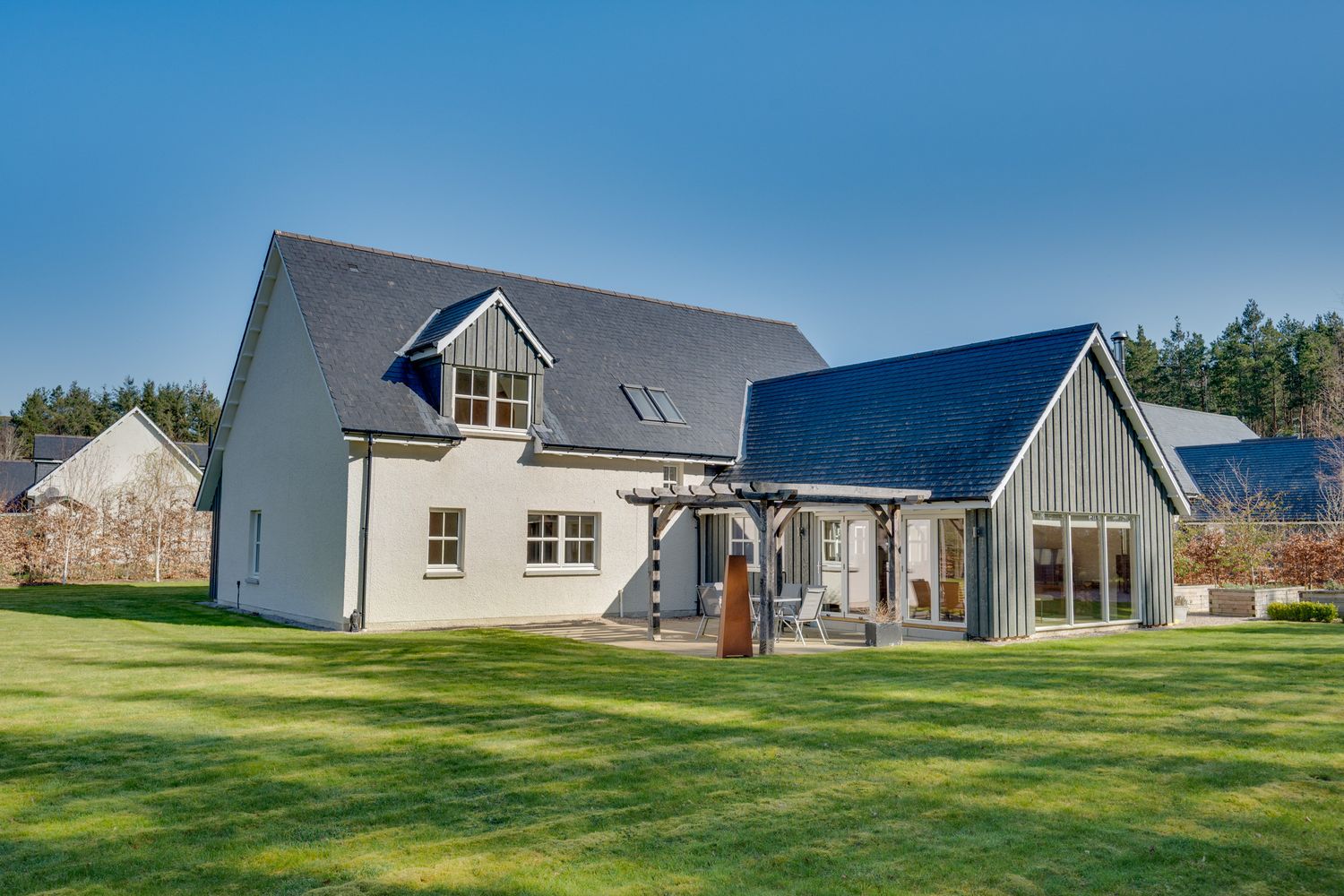 Modern two-story farmhouse with gray siding and blue accents features dormer windows and spacious lawn on sunny day.