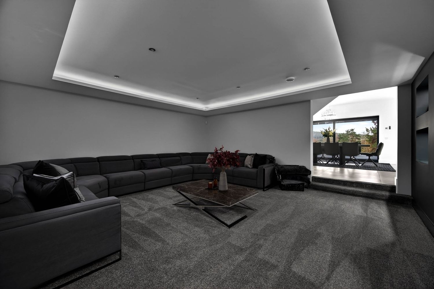 Modern living room with gray sectional sofa, tray ceiling with ambient lighting, and large window overlooking city views.