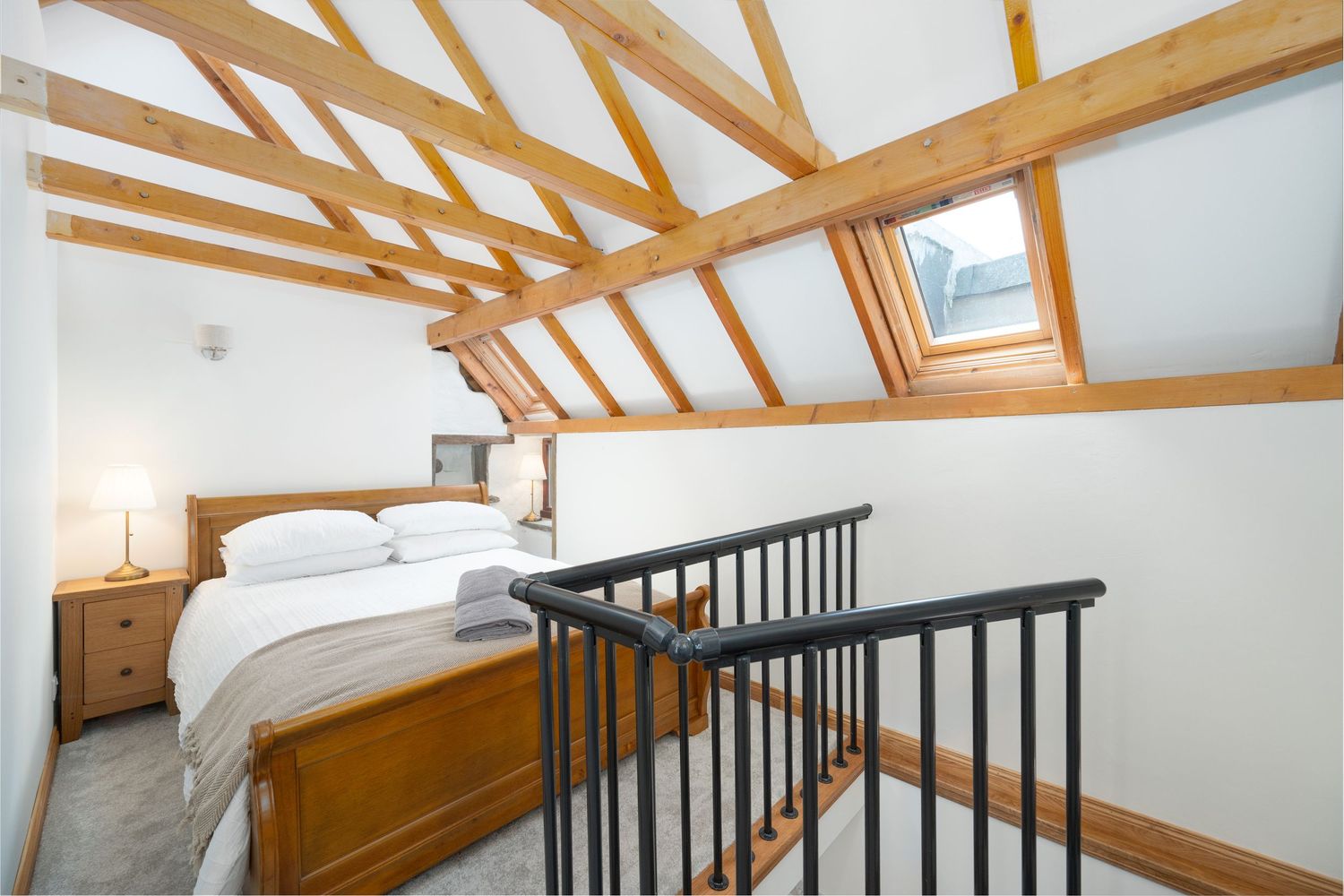 Cozy loft bedroom with exposed wooden beams and skylights featuring a white bed and black metal railing.