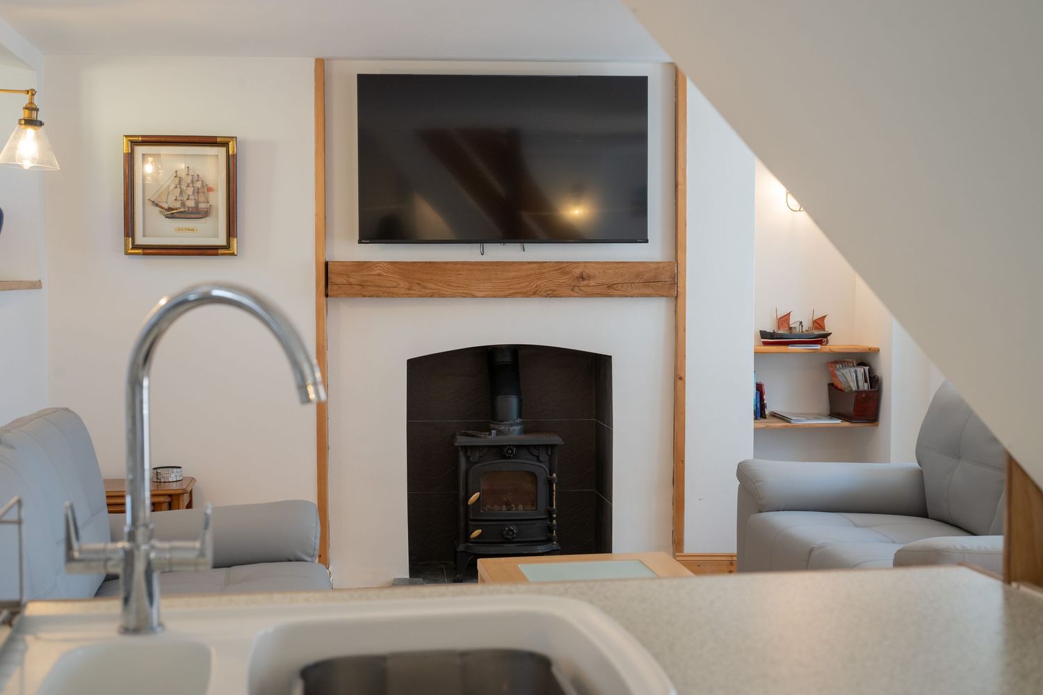 Modern kitchen view with sink, wood-burning stove, mounted TV, and gray armchair in a cozy living space.