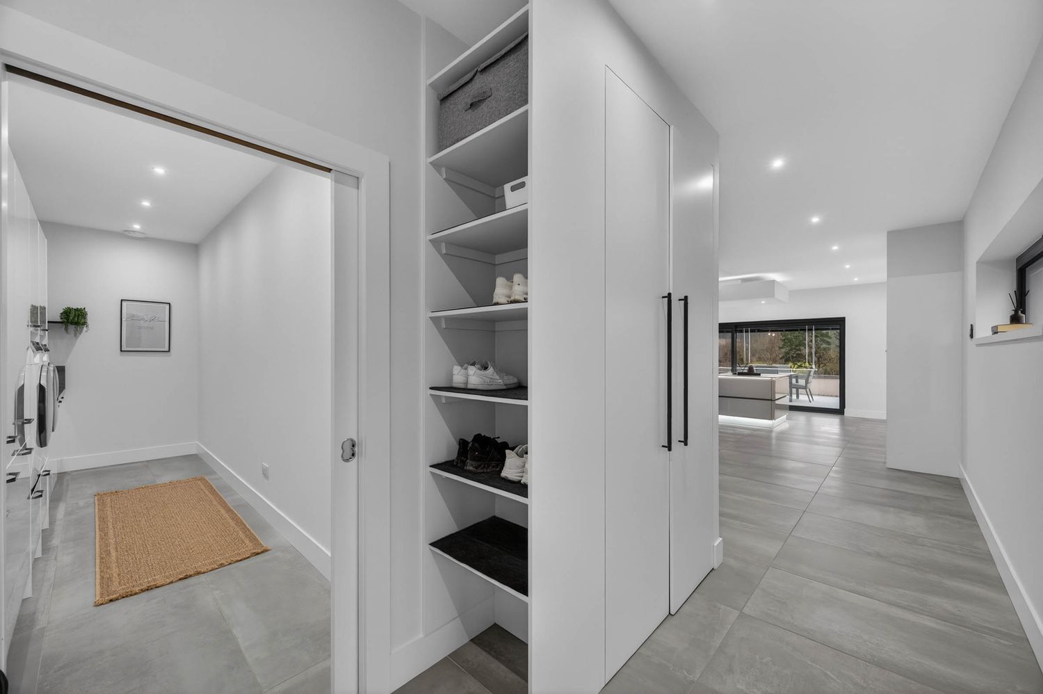 Modern white hallway with built-in shelving, grey flooring, and a tan doormat leading to an open concept living space.