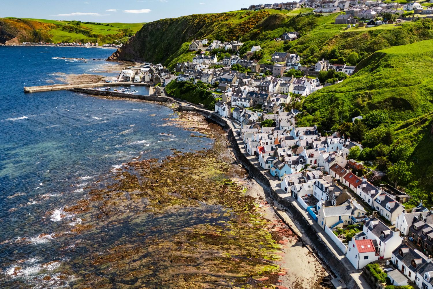 Aerial view of coastal town with white buildings perched along curved rocky shoreline and blue waters.