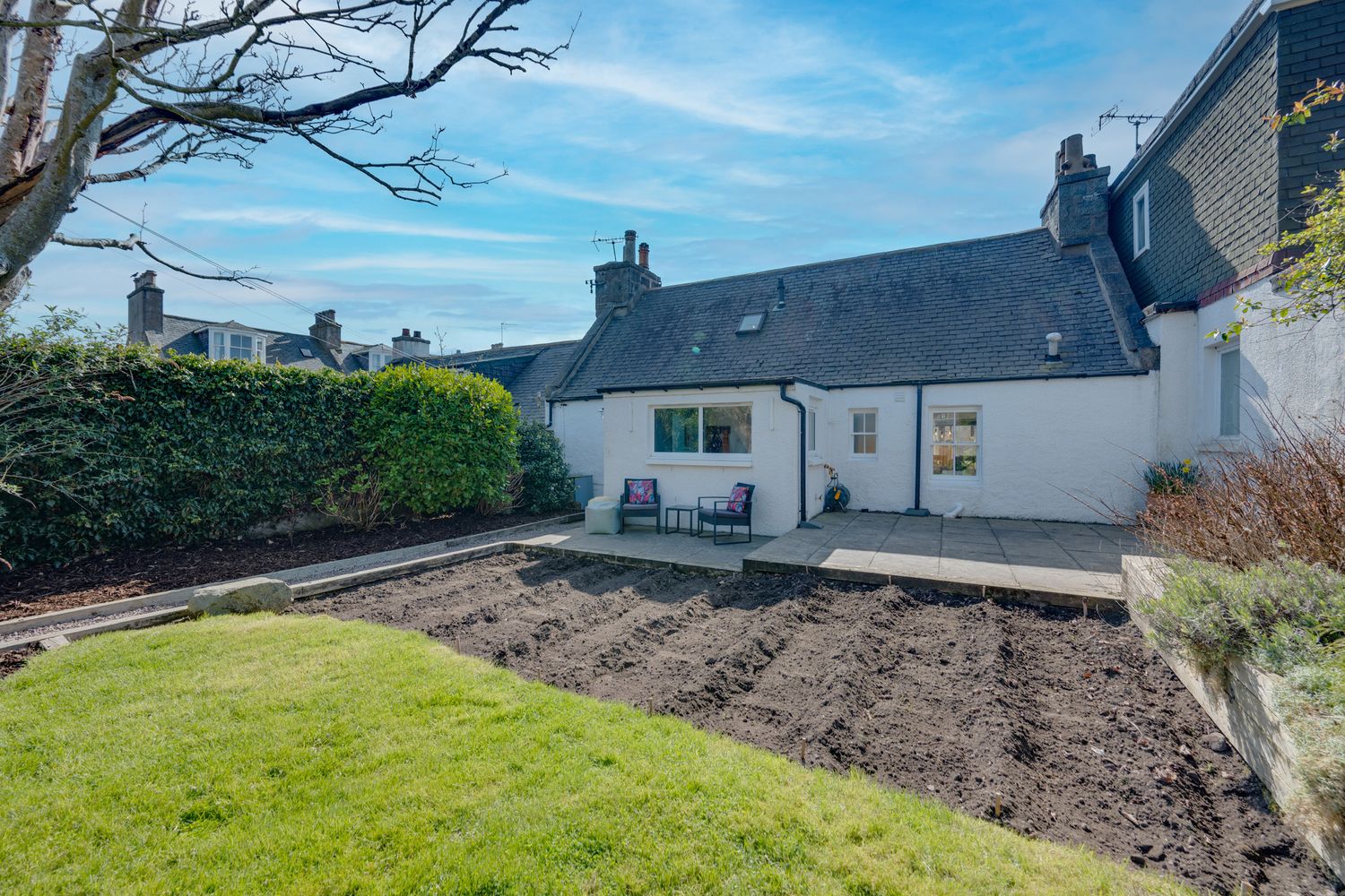 White cottage house with newly tilled garden bed next to green lawn on sunny day with blue sky.