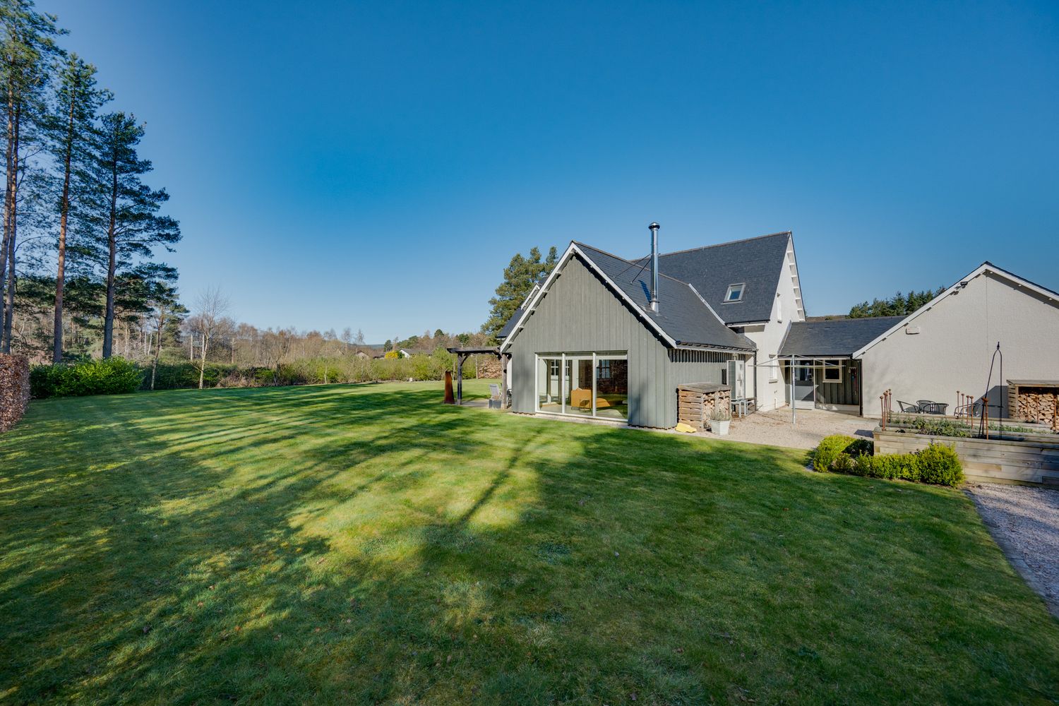 Modern gray house with dormer windows sits on expansive green lawn surrounded by tall pine trees on sunny day.