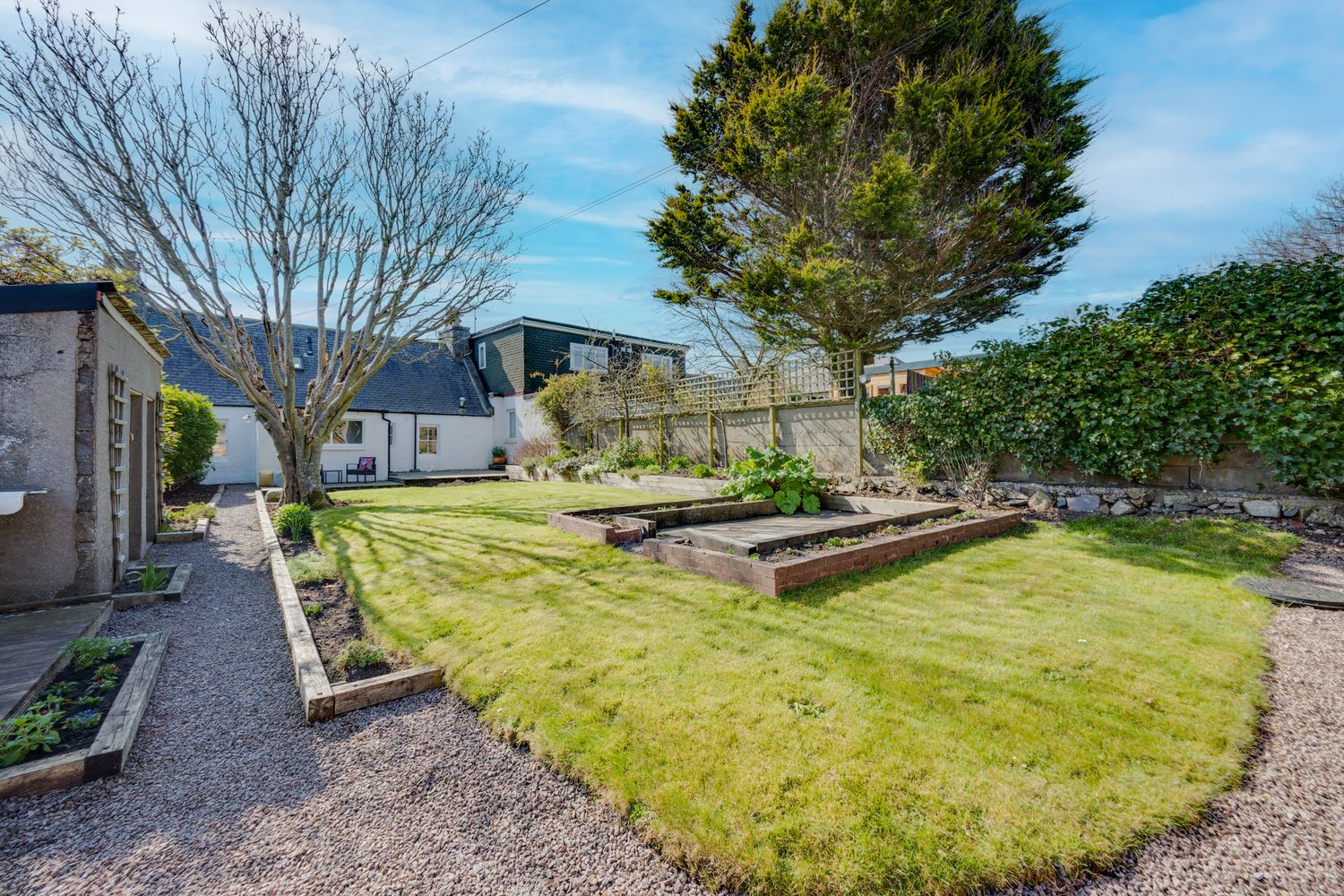 A well-maintained backyard garden with raised beds, a manicured lawn, and evergreen trees under blue skies.