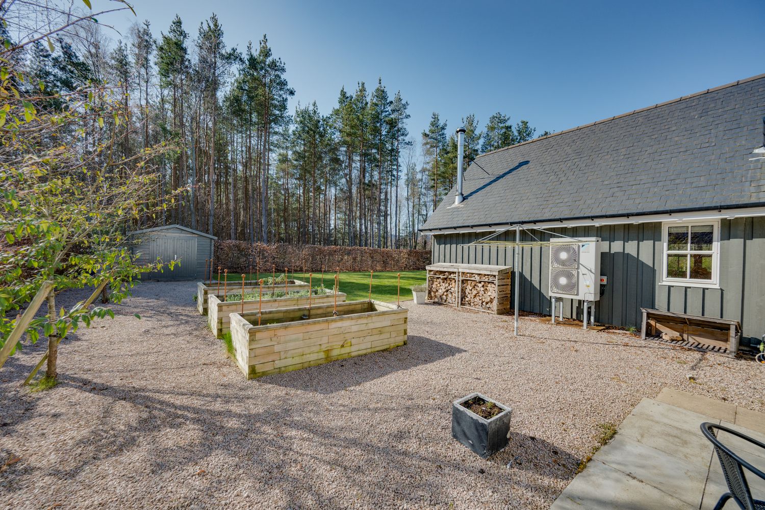 Modern house exterior with raised garden beds and gravel landscaping surrounded by tall pine trees.