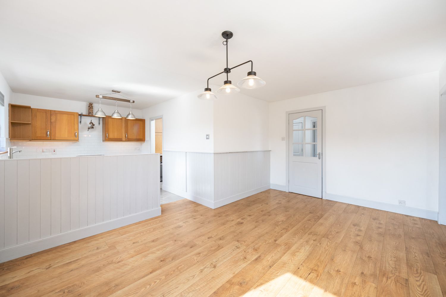 Empty residential room with hardwood floors, white walls, wainscoting, and wooden kitchen cabinets visible in background.