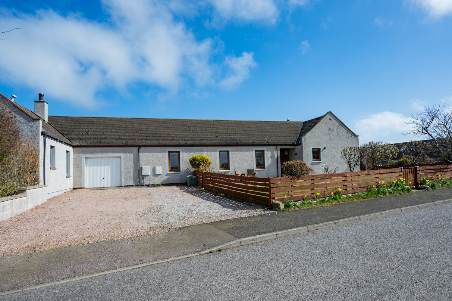 Modern white bungalow with attached garage and wooden deck on a sloped driveway with gravel landscaping and mountain views.