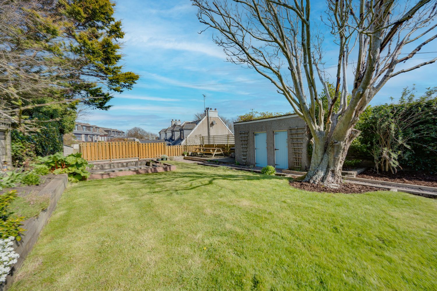 Well-maintained backyard with lush green lawn, wooden fence, garden shed and mature trees against blue sky.