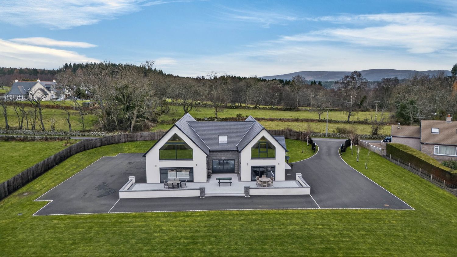 Modern white house with dormer windows nestled in rolling green countryside under blue skies.