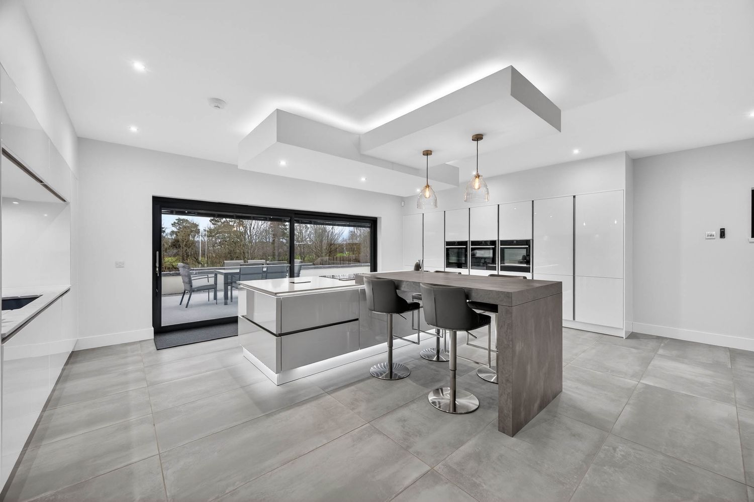 Modern white kitchen and living space with sleek minimalist design, grey flooring, and recessed lighting throughout.