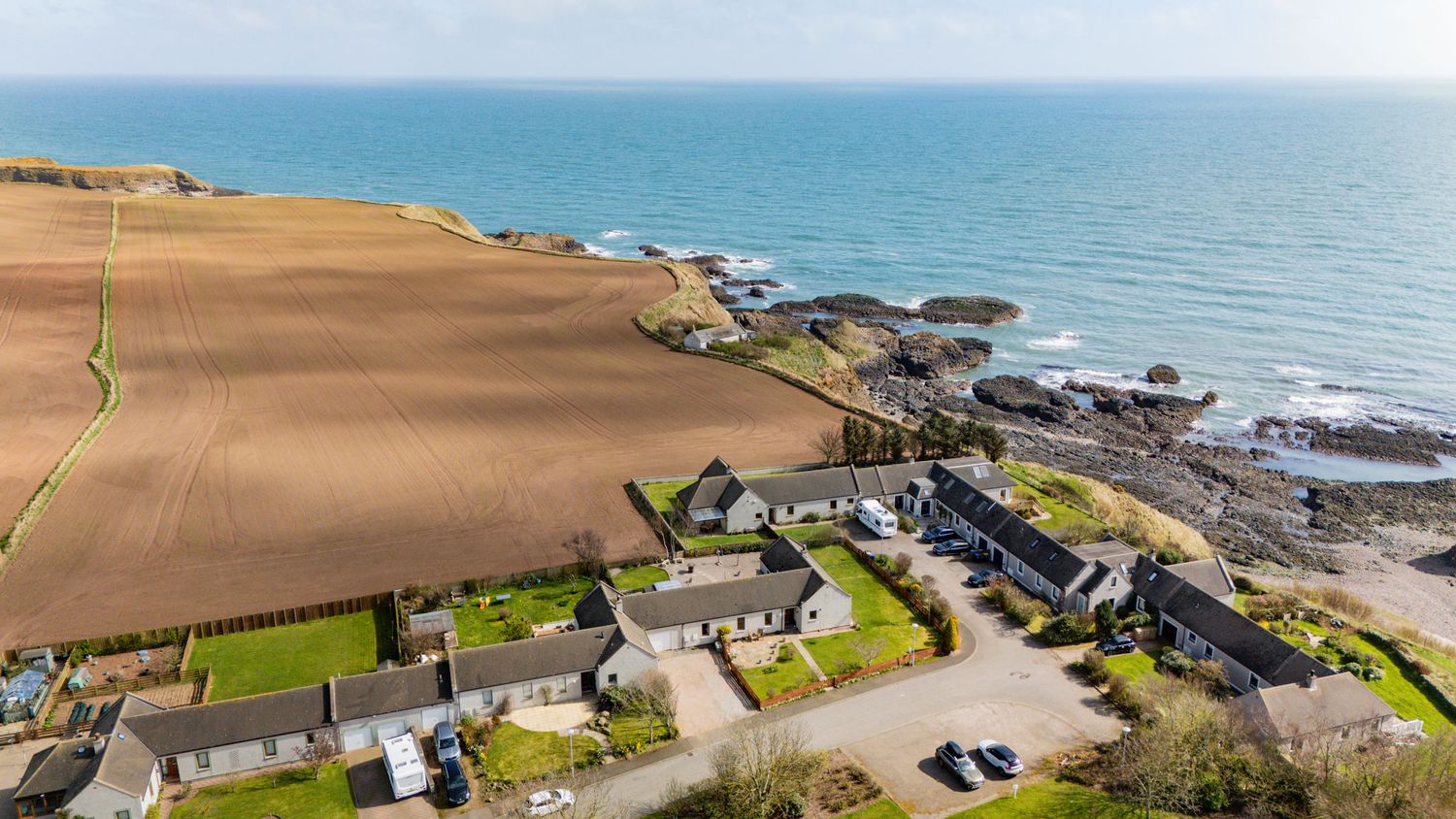 Aerial view of coastal homes and farmland along rocky shoreline with blue ocean waters in the background.