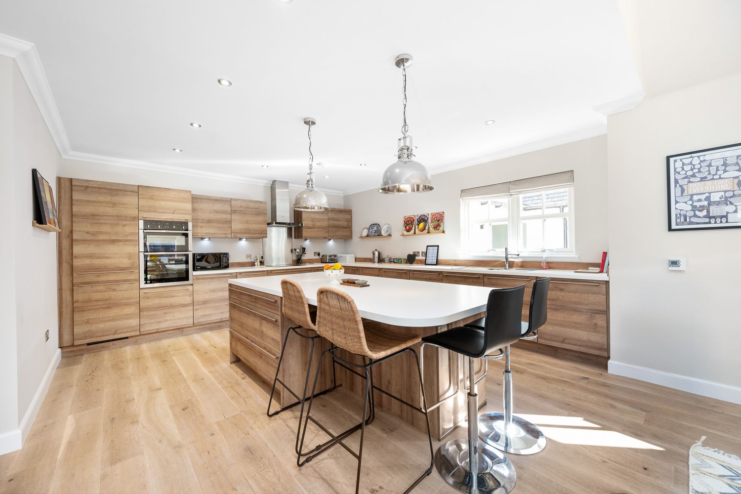 Modern kitchen interior with wooden cabinets, white countertops, and bar stools around a central island with pendant lights.