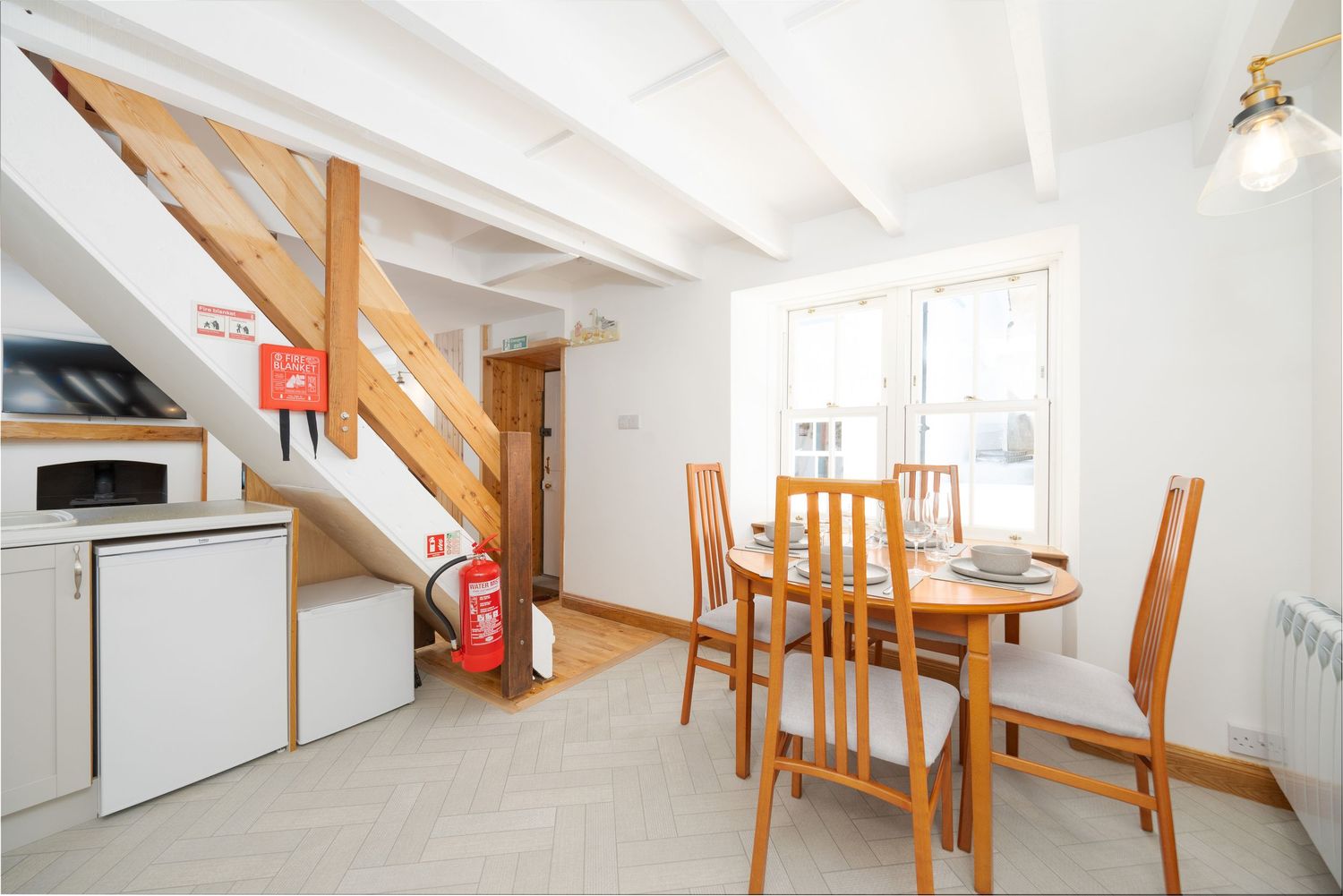 Compact dining area with wooden table and chairs next to white kitchen and wooden staircase.