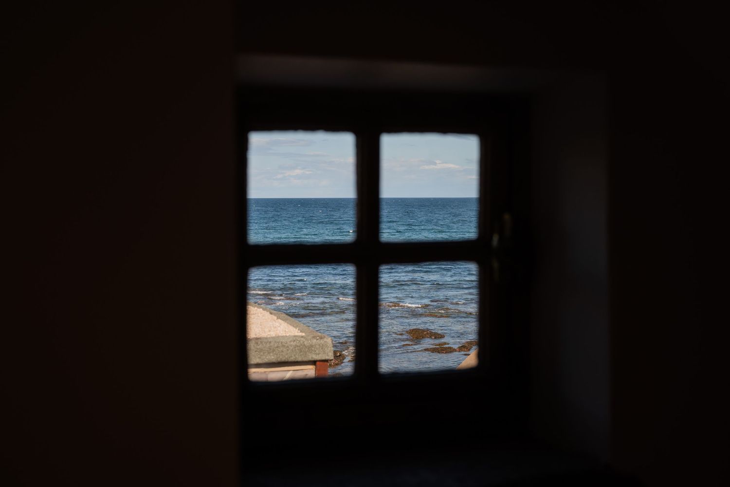 View of a sandy beach and ocean horizon through a dark window frame creating a moody composition.