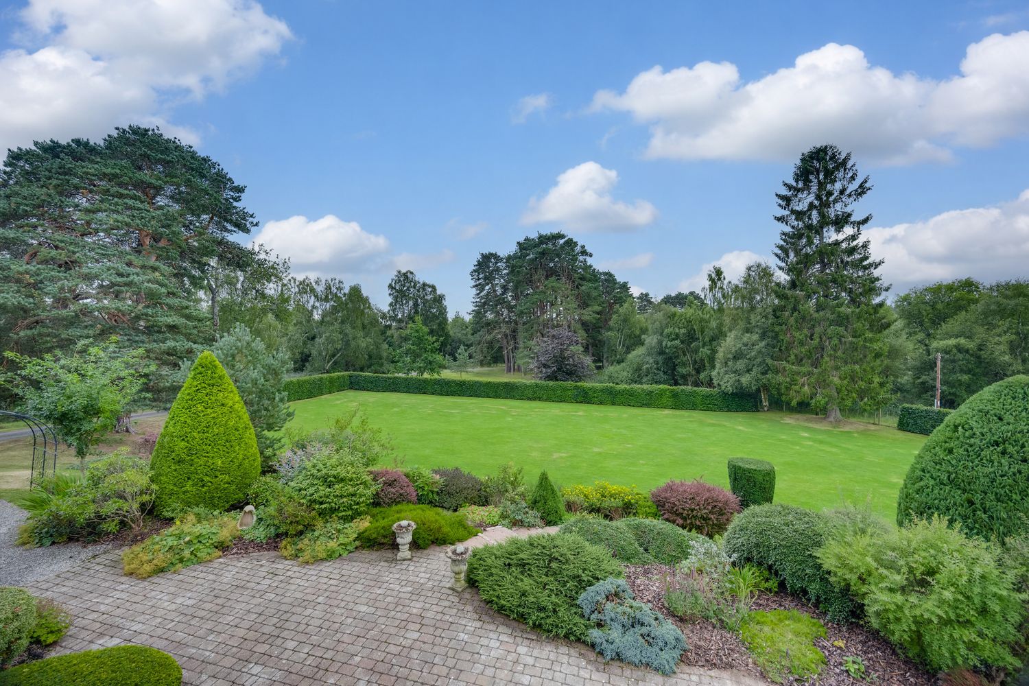 Manicured English garden with lush green lawn, topiary shrubs, and tall trees under a bright blue sky.