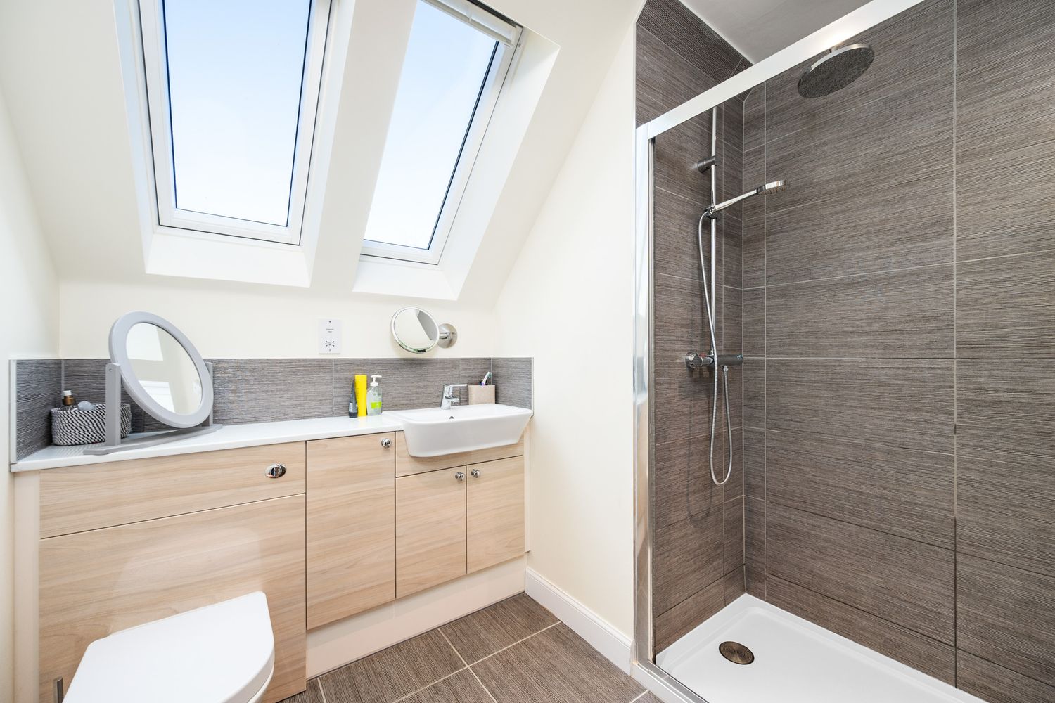 Modern attic bathroom with sloped ceiling featuring wood cabinets, grey tile walls, and glass walk-in shower enclosure.