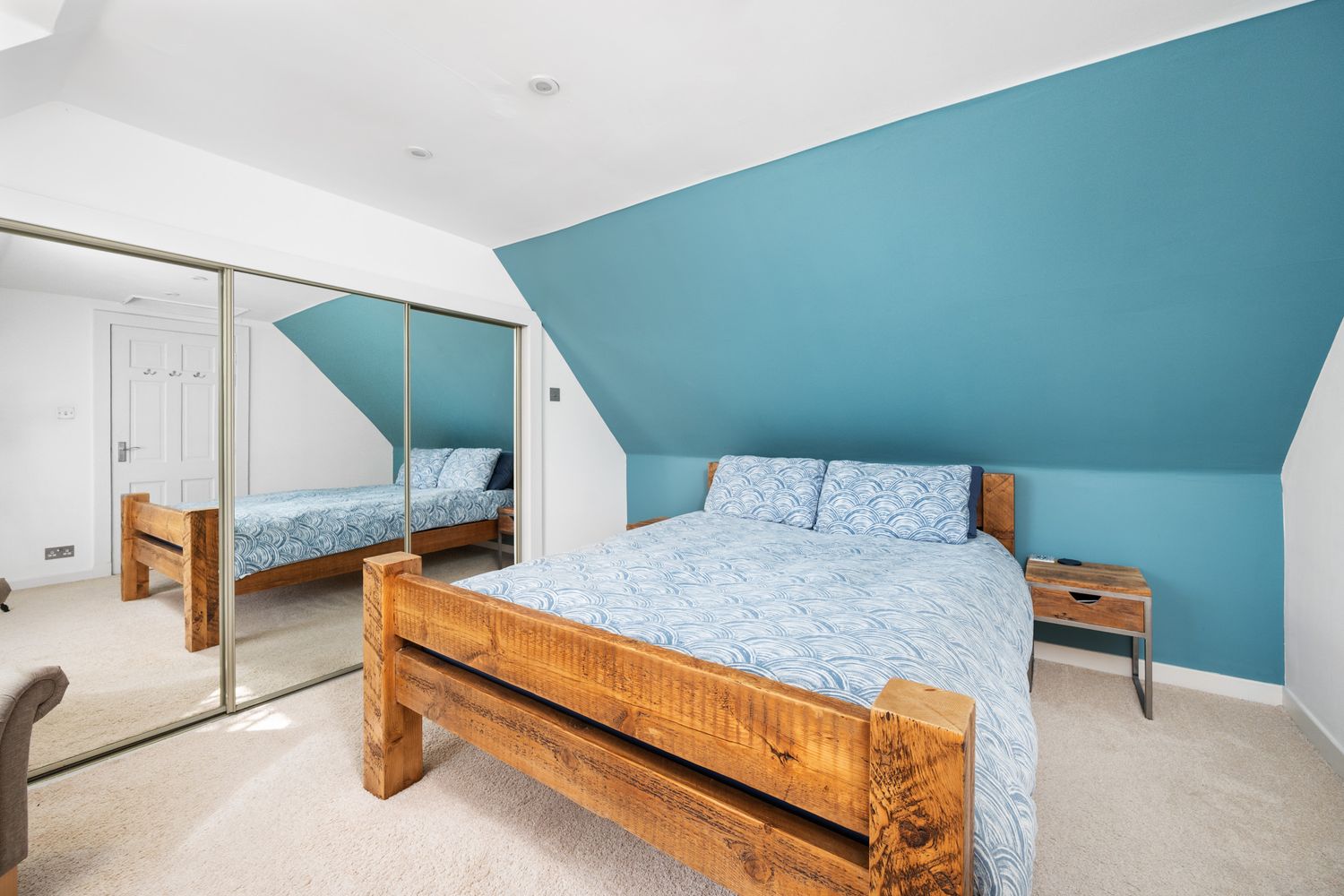Airy bedroom with sloped ceiling featuring a wooden bed frame, blue accent wall, and full-length mirror.