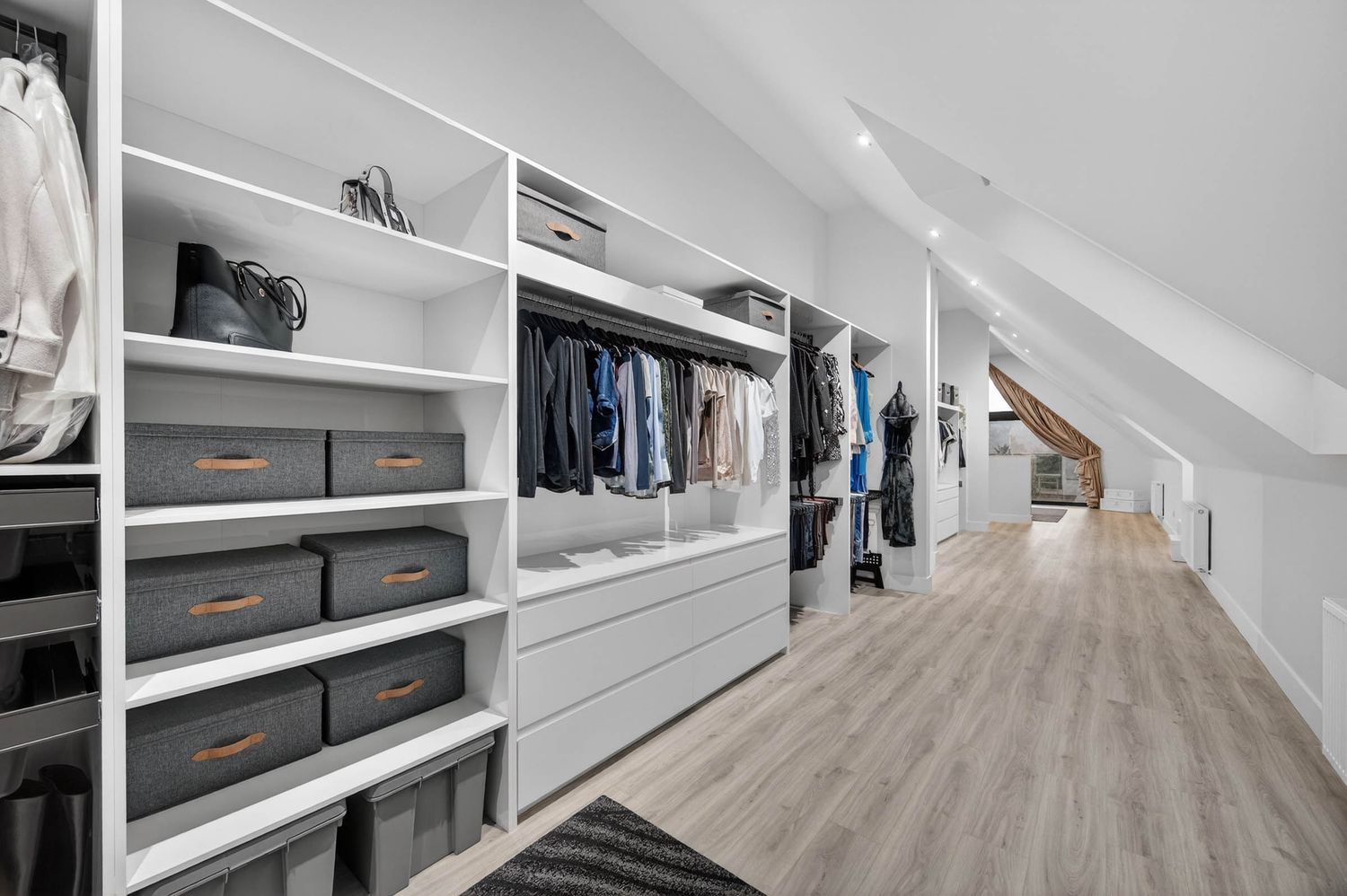 Modern white walk-in closet with sloped ceiling features built-in shelving and hardwood flooring.