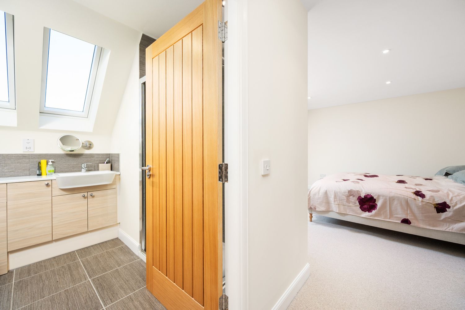 Interior view showing wooden door between modern bathroom with tiled floor and bright bedroom with floral bedding.