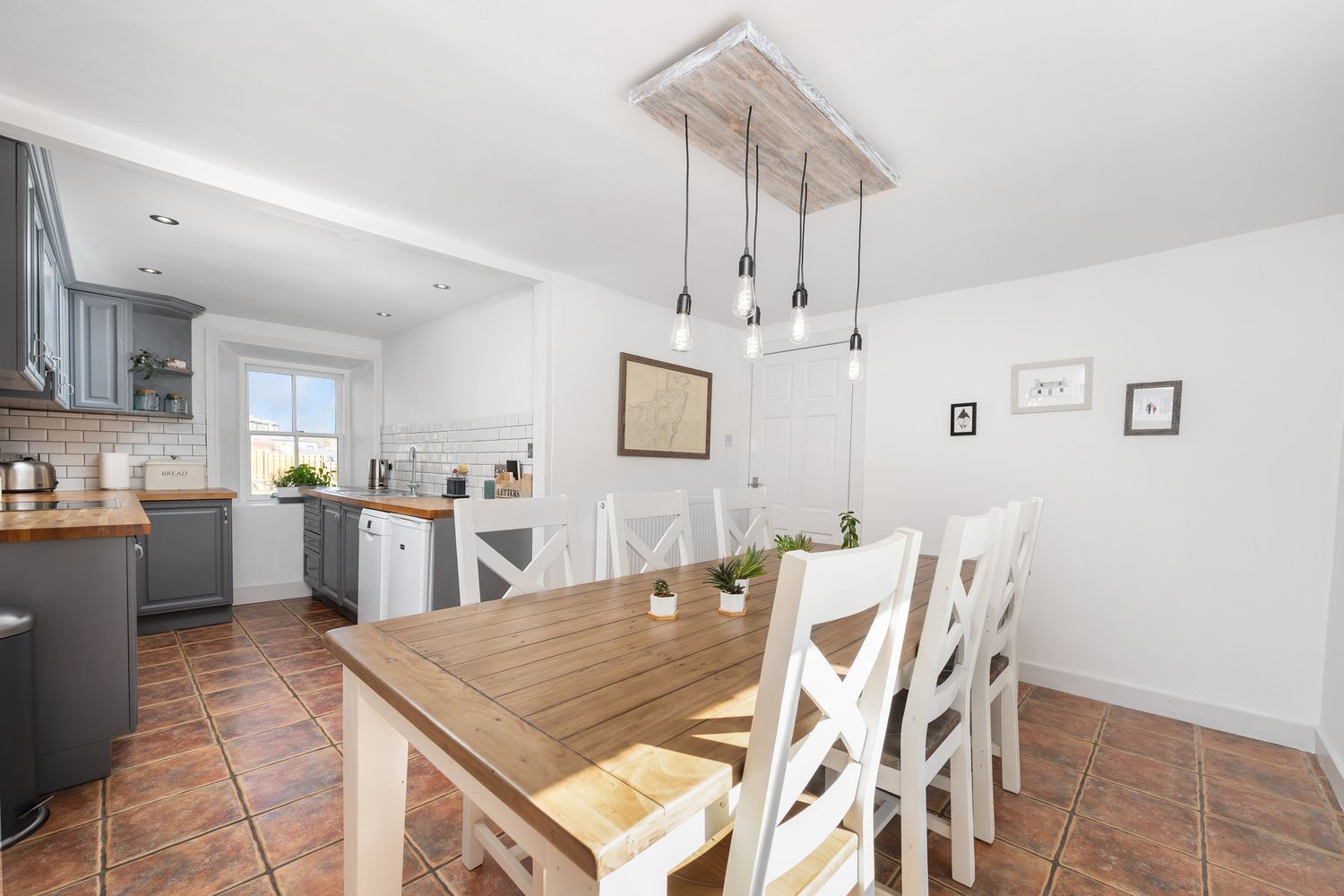 Modern kitchen and dining area with terracotta floors, white walls, and wooden dining table with cross-back chairs.