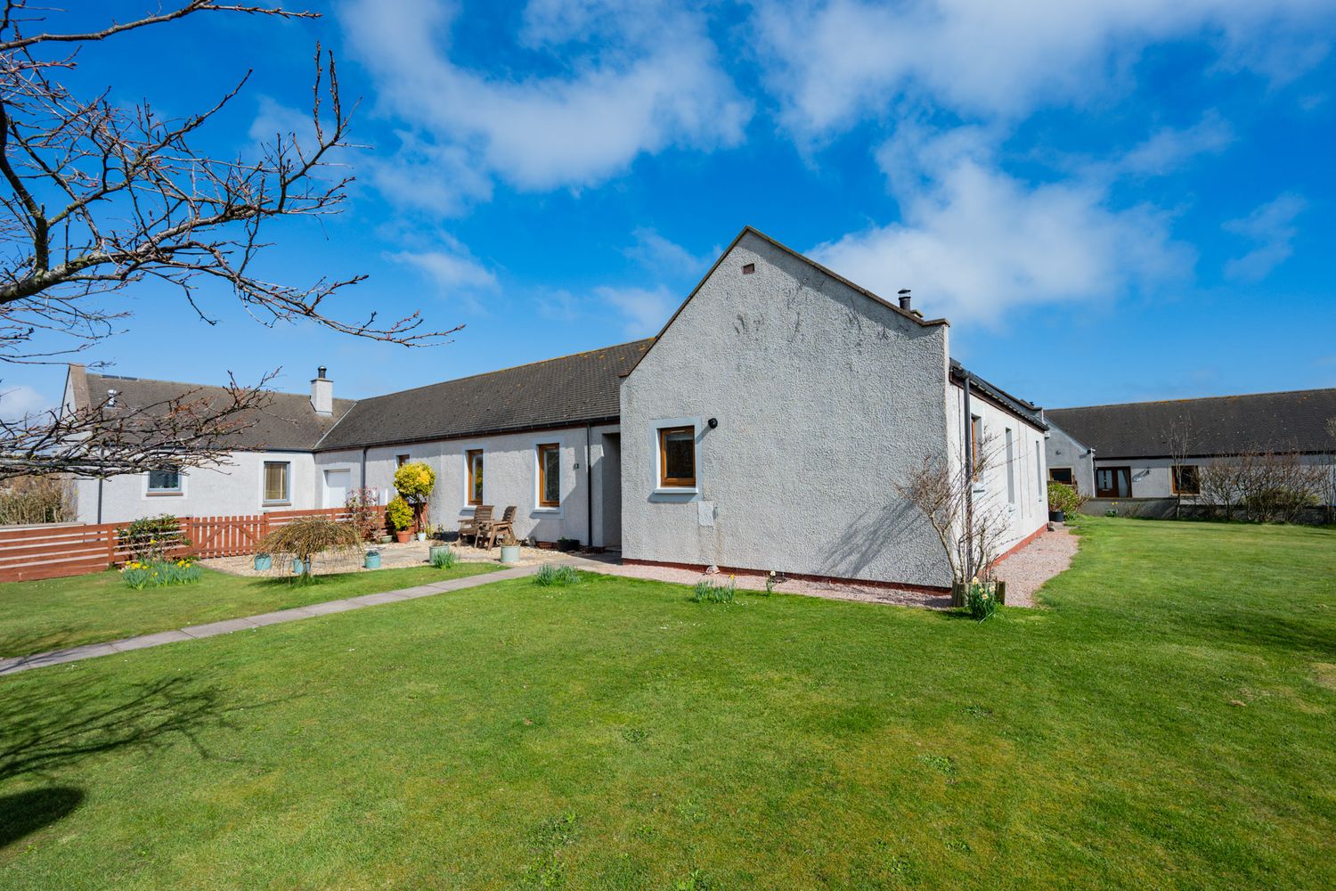 Single-story white bungalow with spacious green lawn and grey exterior set against bright blue sky and clouds.