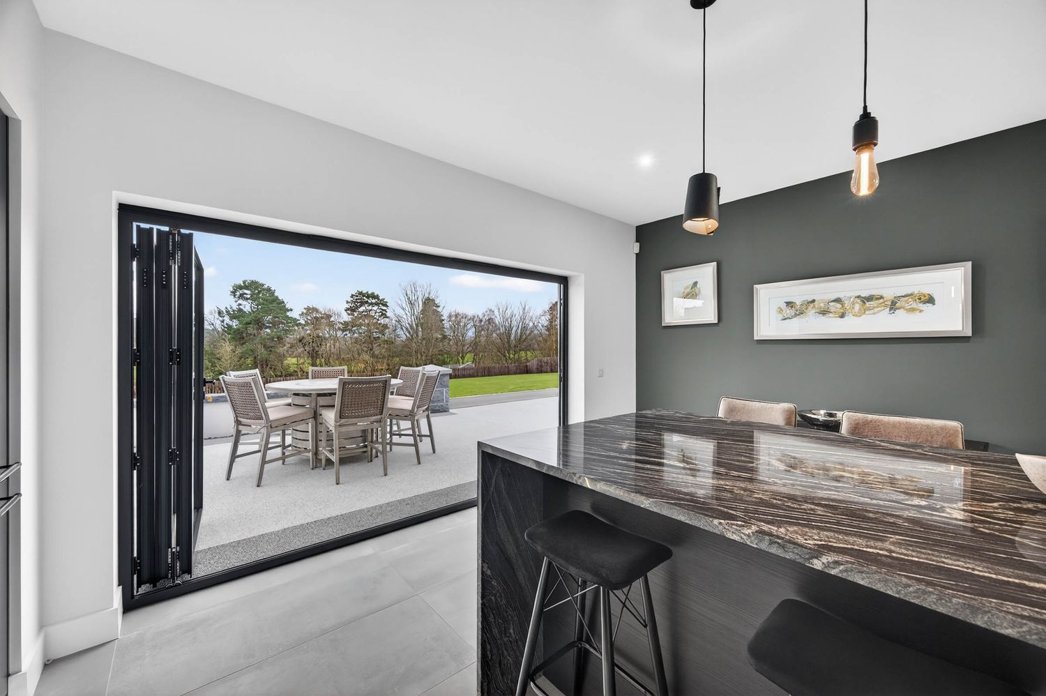 Modern kitchen with dark accent wall opens to patio through folding glass doors, featuring marble countertop and pendant lights.
