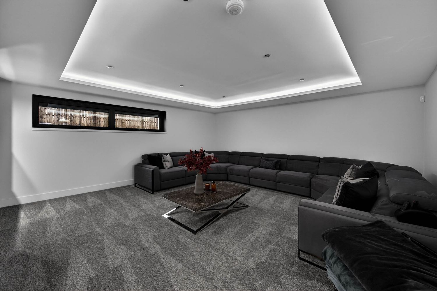 Modern basement living room with dark sectional sofa, glass coffee table and recessed lighting in monochromatic style.
