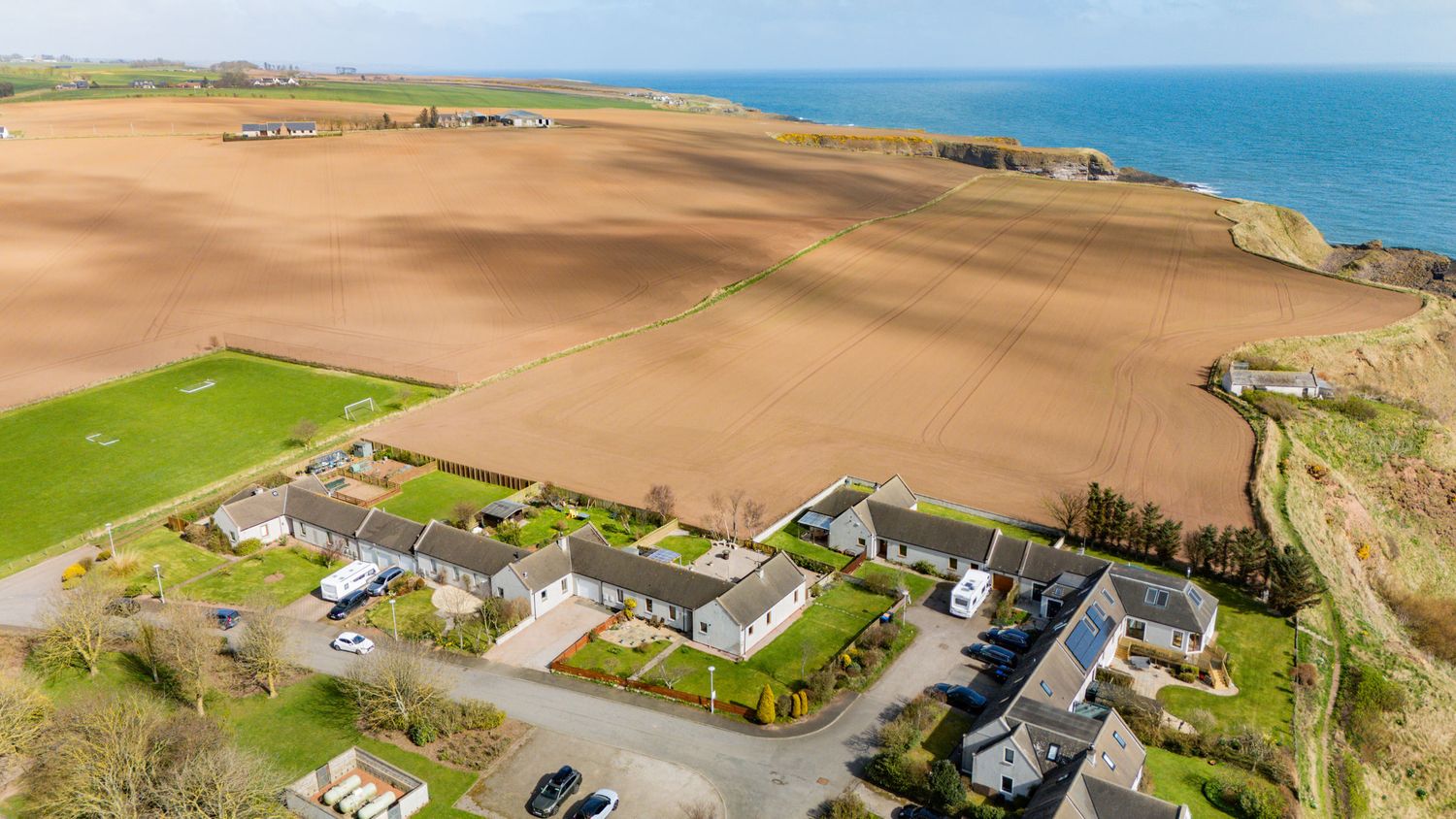 Aerial view of coastal homes and farm fields along dramatic ocean cliffs on a sunny day.