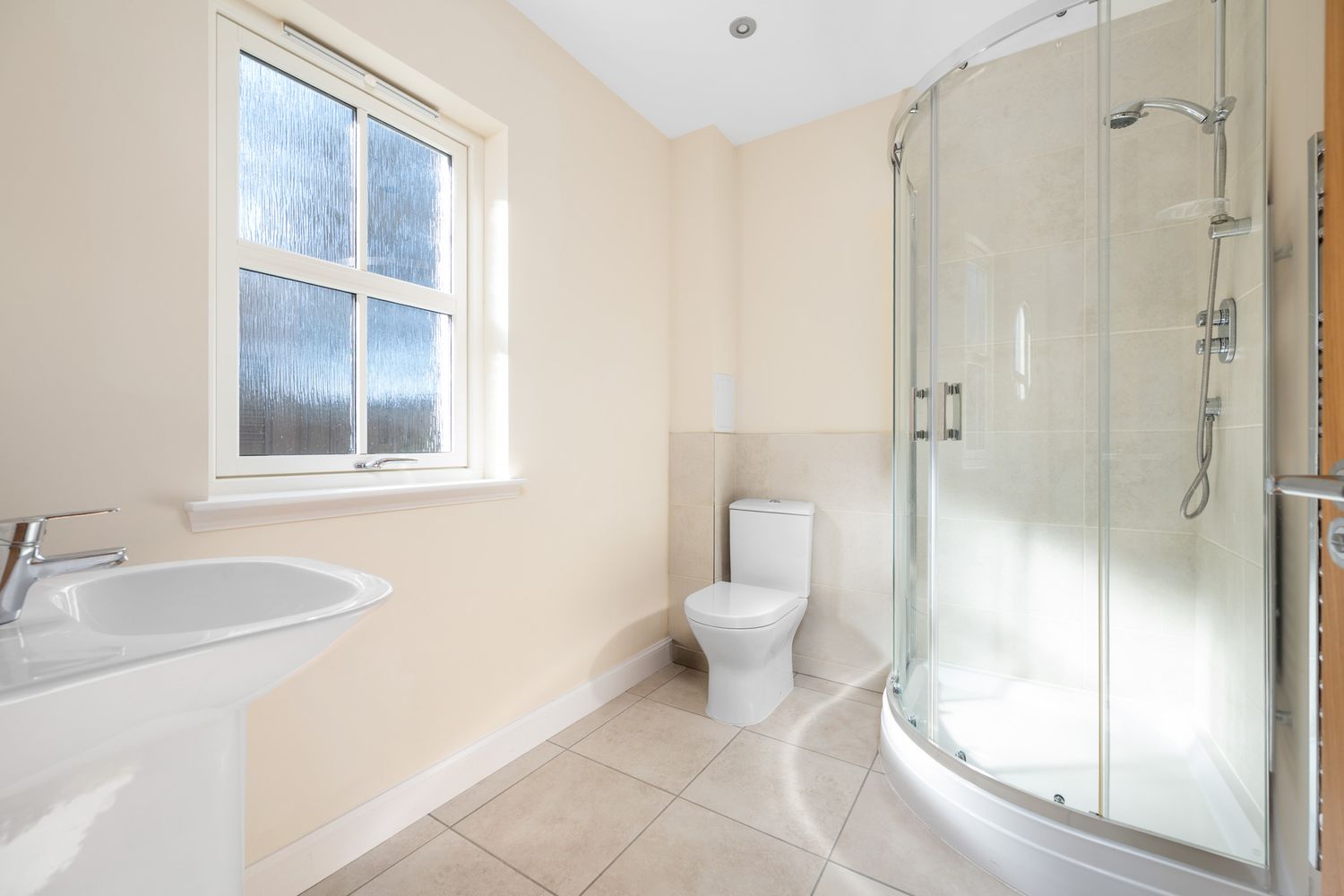 Modern white bathroom with curved glass shower enclosure, pedestal sink, and natural light from window.