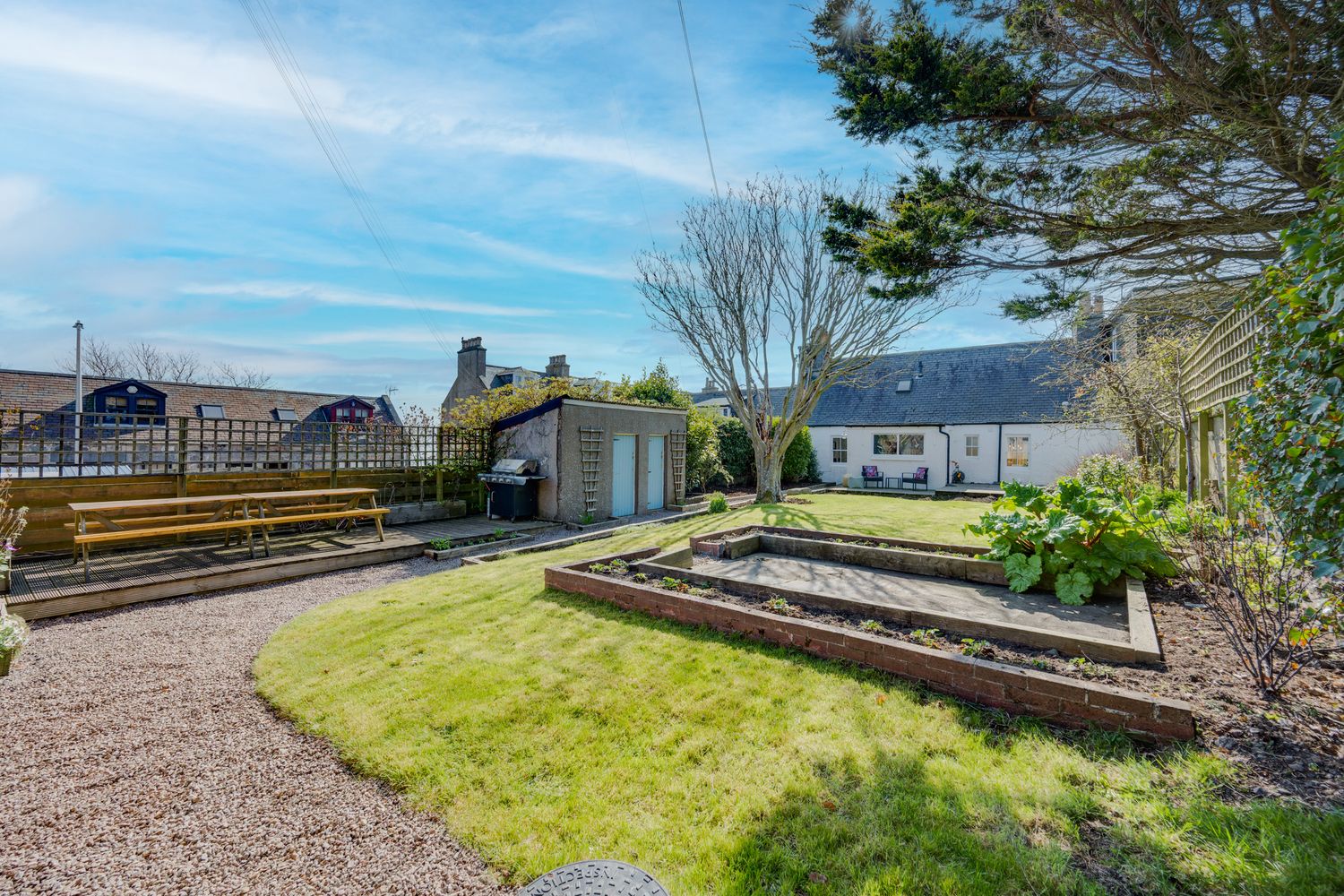 A backyard garden with raised beds, gravel path, and well-maintained lawn under blue skies.