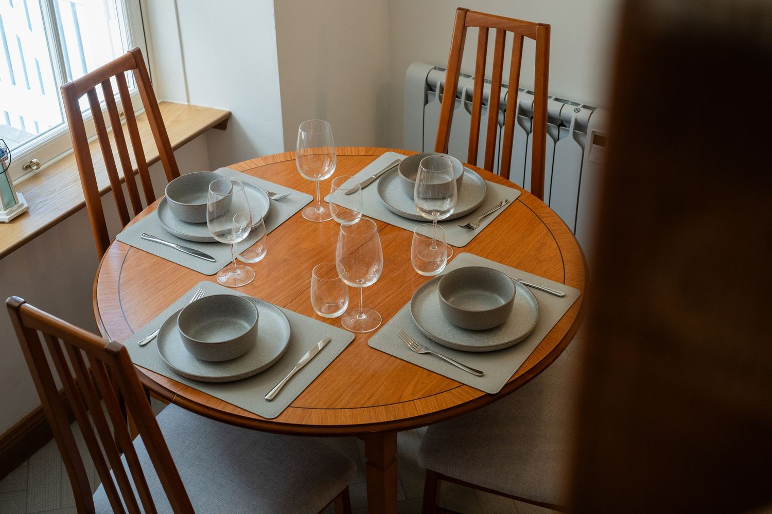 Round wooden dining table set with plates, wine glasses and cutlery for four people in a home setting.