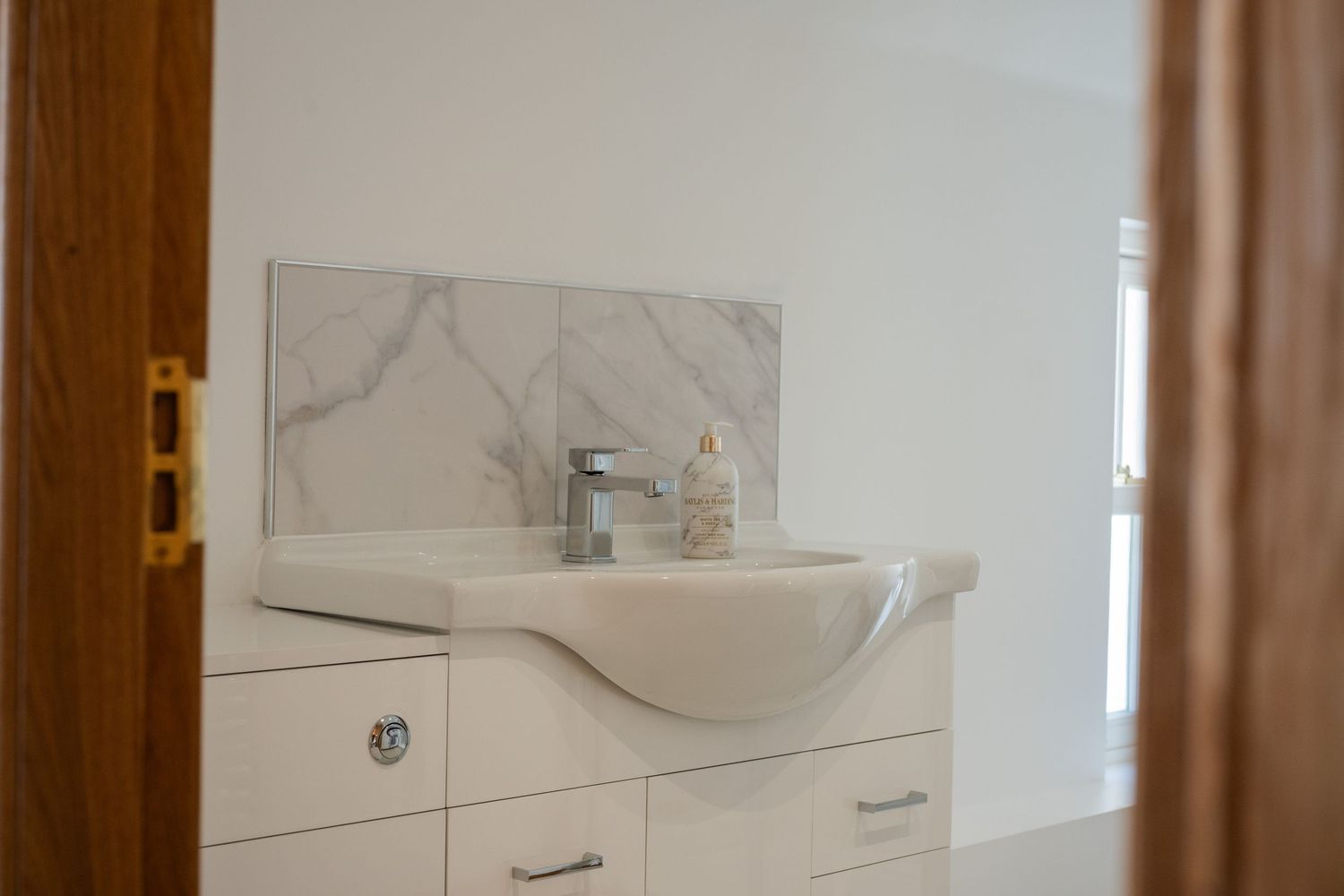 Modern white bathroom vanity with curved sink and chrome faucet against marble-style backsplash in bright interior.