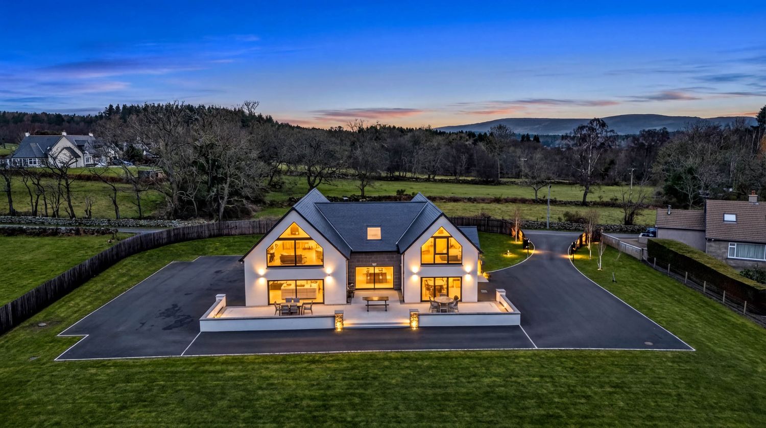 Modern A-frame house with large glass windows glowing warmly at dusk on a spacious green lawn with mountains in background.