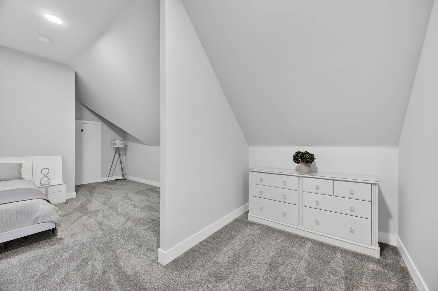 A white interior room with sloped ceiling, grey carpeting, and a small window shows modern minimalist design elements.