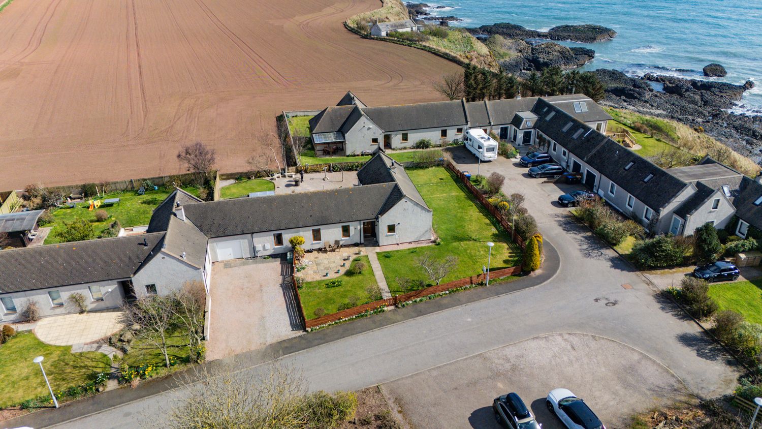 Aerial drone view of coastal homes and buildings along a rocky shoreline with farmland and parking area in Scotland.