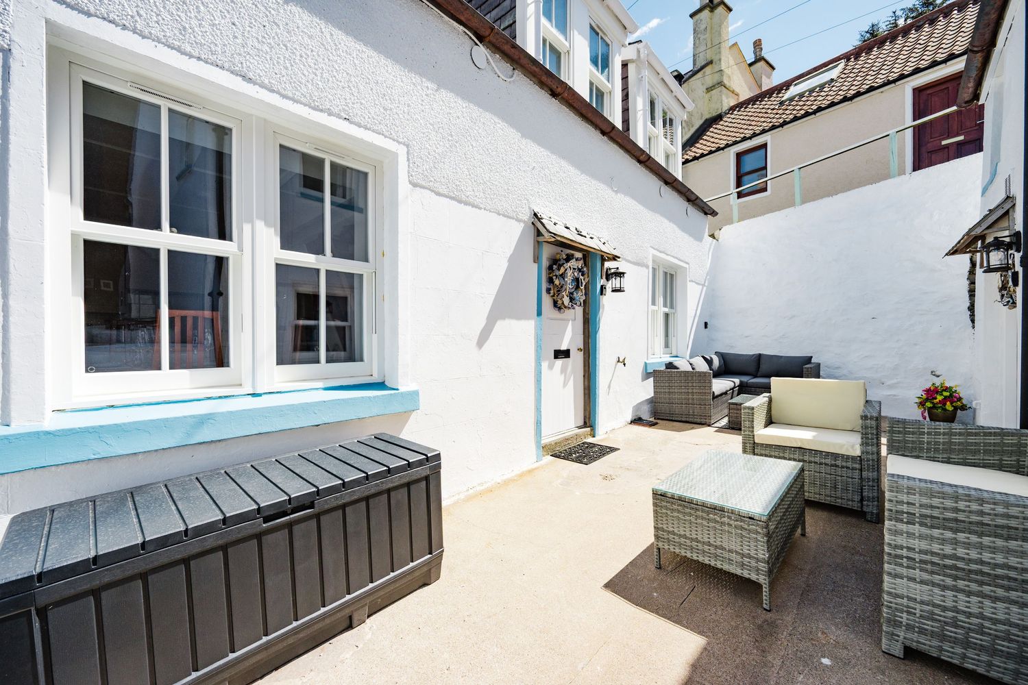 A cozy patio terrace with a black hot tub, grey rattan furniture, and white walls featuring blue-trimmed windows.