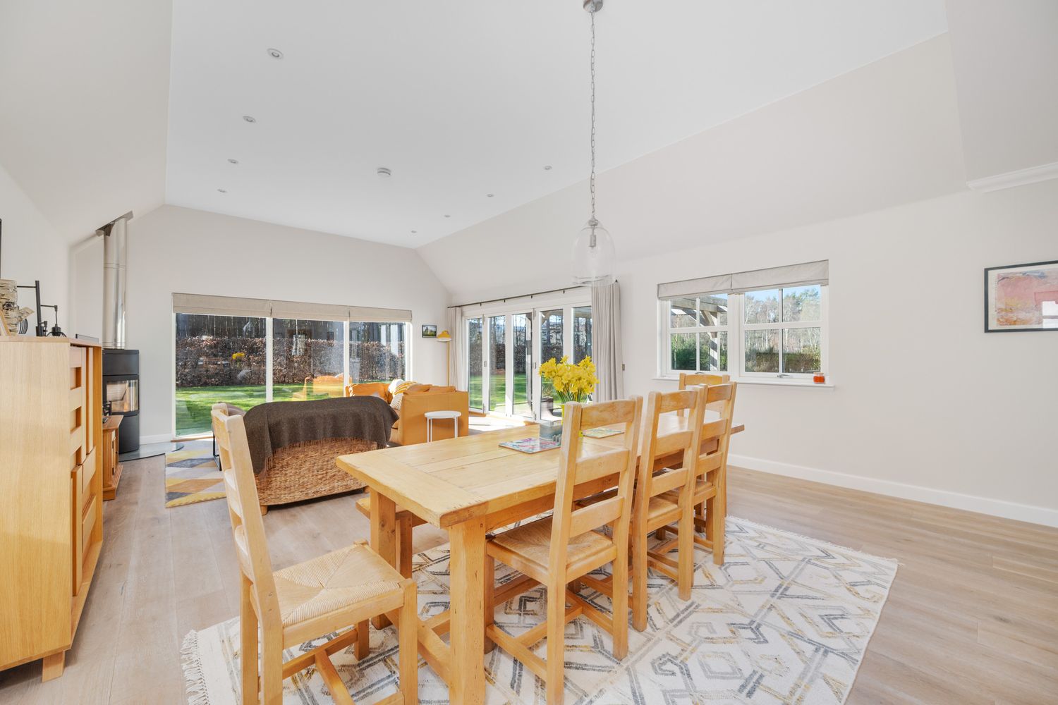 Modern dining room with wooden table set, gray area rug, and large windows overlooking a garden.