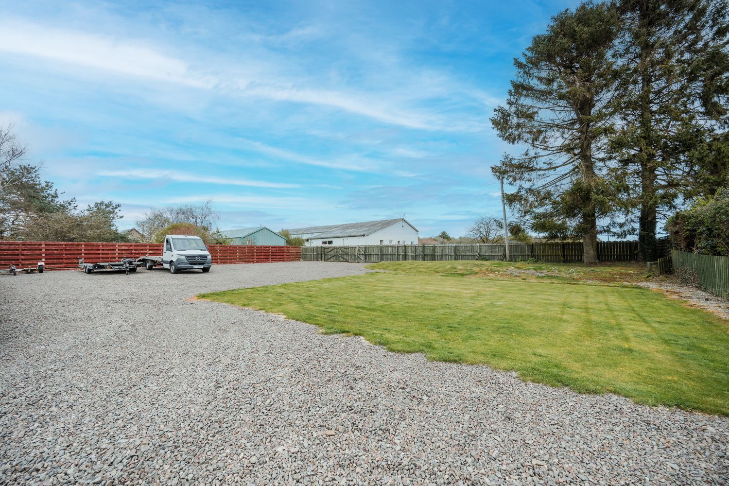 A gravel parking area next to a green lawn with trees and fencing under bright blue skies.