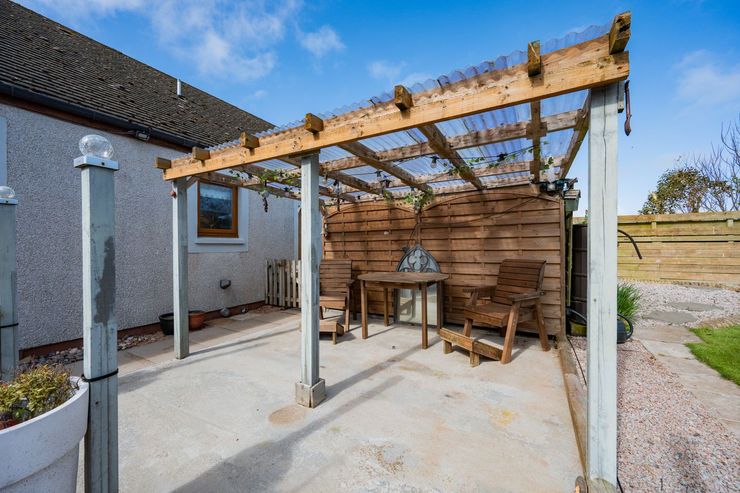 A wooden pergola with outdoor seating and grill area attached to a white stucco house with concrete patio.