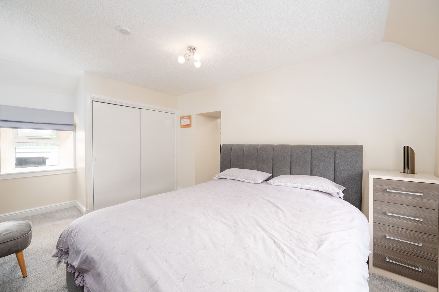 Modern bedroom with grey upholstered headboard, white bedding, grey dresser and grey accent chair near window.